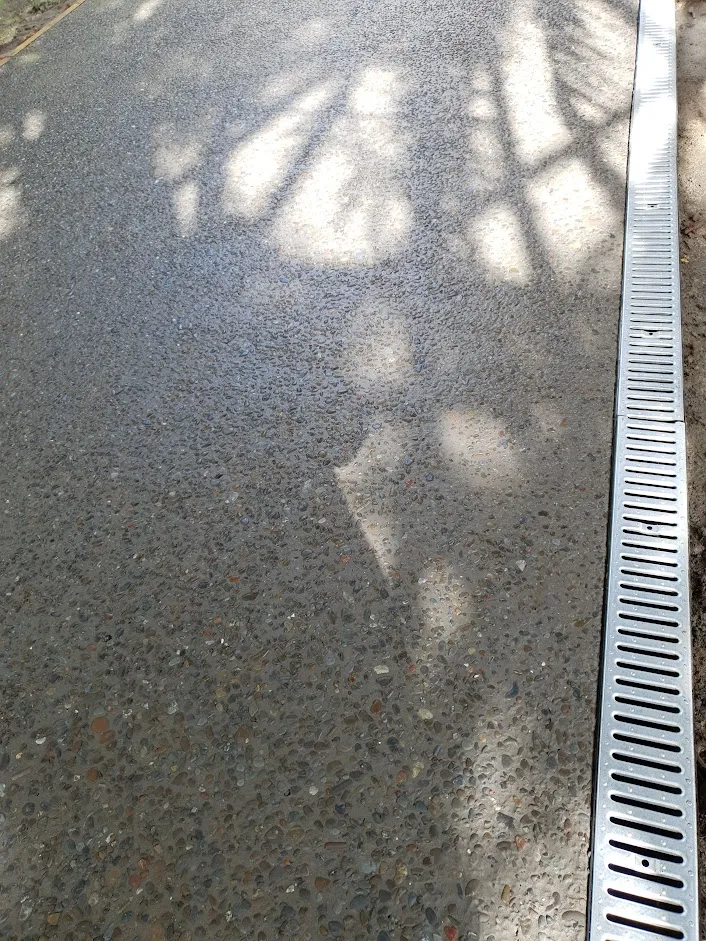 Pebbled concrete pathway with shadows of tree branches and a metal drainage grate on the right side.